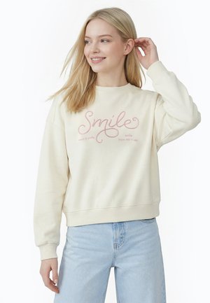 Ung kvinde med blondt hår iført en cremefarvet sweatshirt med teksten "Smile" og lyseblå jeans, der rører ved sit øre og smiler.