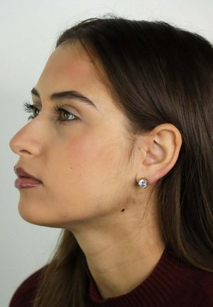 Jonge vrouw met lang donker haar, gekleed in een donkerrood shirt en studs oorbellen, kijkt naar links tegen een effen achtergrond.