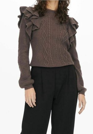 Femme portant un pull en maille torsadée marron avec des épaules à volants et un pantalon noir taille haute, debout devant un fond blanc.