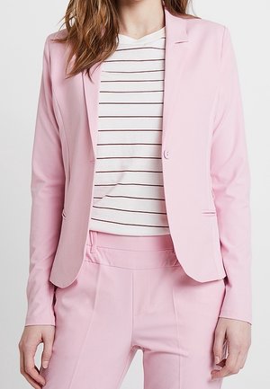 Femme portant un blazer rose clair cintré et un pantalon assorti sur une chemise blanche à fines rayures horizontales noires.