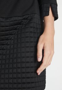 Svart klänning med en slät överdel och en texturerad, kviltad nedre del. Långa ärmar med en fyrkantig manschettdetalj.