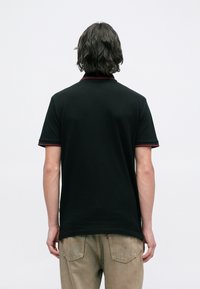 Person with medium-length dark hair wearing a black short-sleeve polo shirt with red trim and beige pants, facing away on a white background.