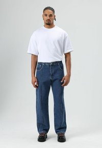 T-shirt blanc oversize, jean bleu en denim avec surpiqûres contrastantes, coupe décontractée, et chaussures noires épaisses à lacets. Fond uni.