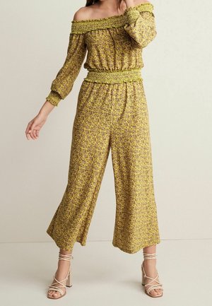 Femme portant une combinaison longue jaune à fleurs, épaules dénudées, manches longues, taille smockée, et des sandales beiges à talons hauts à brides, debout sur un fond uni.