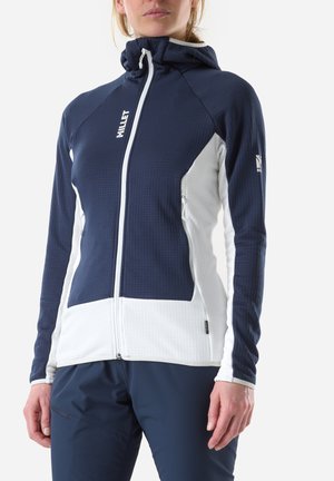 Woman wearing navy and white zip-up hooded jacket with branding on chest and sleeve, paired with navy pants, standing against white background.