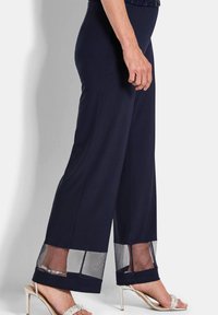 Vrouw in een marineblauwe broek met sheer mesh panels aan de zoom en zilveren hoge hakken, staand tegen een lichte achtergrond.