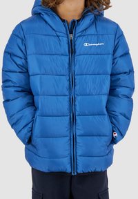 Blå puffjacka med huva, framzip, och elastiska ärmar. Har horisontell sömnad och en liten logotyp på vänster bröst. Mjuk textur.