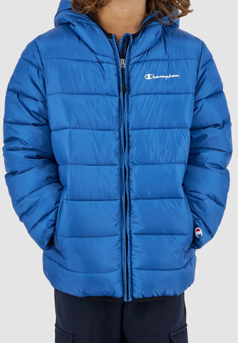 Blå puffjacka med huva, framzip, och elastiska ärmar. Har horisontell sömnad och en liten logotyp på vänster bröst. Mjuk textur.