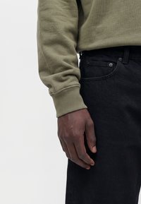 Main gauche d'une personne portant une chemise à manches longues olive clair et un jean noir visible sur un fond blanc uni.