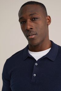 Polotriko tmavě modré barvy z pletené látky, s klasickým límečkem, dvouknoflíkovým zapínáním a strukturovaným povrchem. Nositelná přes bílé tričko.