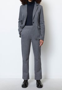 Grijs geruite pak met een op maat gemaakte blazer en bijpassende broek. De stof heeft een subtiele textuur. Gepaard met een zwarte coltrui en laarzen.