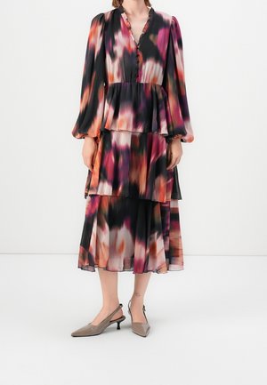 Vrouw die een jurk draagt met lange mouwen, meerdere lagen, met zwarte, roze, oranje en witte abstracte patronen en beige slingbacks met lage hak.