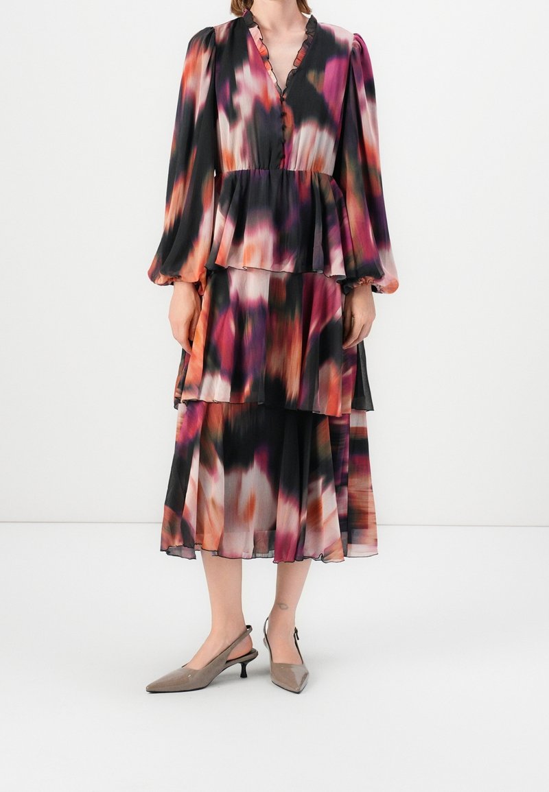 Femme portant une robe à manches longues et multi-couches avec des motifs abstraits noirs, roses, orange et blancs, et des chaussures slingback beiges à talon bas.