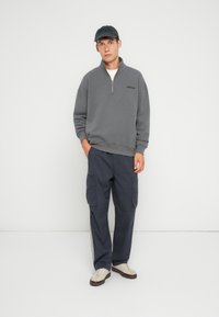 Sudadera gris con cremallera combinada con pantalones cargo azul marino, que cuentan con bolsillos laterales y un corte relajado. Se lleva con una gorra negra y zapatillas beige.