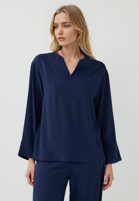 Dunkelblaues Langarmshirt mit V-Ausschnitt und weit geschnittenen Ärmeln. Hergestellt aus einem weichen Stoff mit subtiler Textur und strukturiertem Vorderteil.