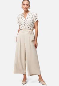 Beige wijde broek met een knoopriemdetail, gedragen met een witte top met een subtiele bloemenprint in zwart en goud. Gladde textuur.