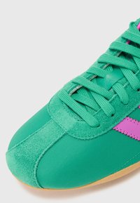 Zeleni športni copat iz semiša in najlona, ki se ponaša z teksturirano zeleno zgornjico, roza poudarjeno črto in gumijastim podplatom. Ima vezalni dizajn.