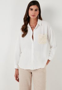 Camisa blanca de botones con un detalle de bolsillo texturizado de crochet y tejido suave y ligero. Mangas largas y corte relajado.