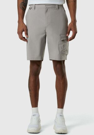 Uomo che indossa pantaloncini cargo grigio chiaro fino al ginocchio con tasca laterale e sneaker bianche su sfondo semplice.