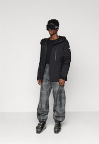 Άντρας φορώντας μαύρο μπουφάν σκι, γκρίζες καρό παντελόνες σκι, μαύρα γάντια σκι, μπότες σκι και μαύρα γυαλιά σκι που στέκεται μπροστά σε απλό φόντο.