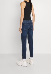 Jeans de mezclilla azul oscuro con un corte ajustado, que cuentan con una cinturilla de altura media y bolsillos traseros. Combinados con zapatillas blancas y de longitud a la altura del tobillo.