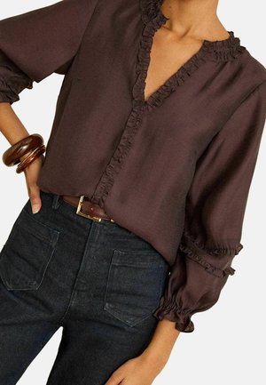 Femme portant un chemisier marron à volants avec manches longues et un jean foncé taille haute, accessoirisée de bracelets marron et d'une ceinture.