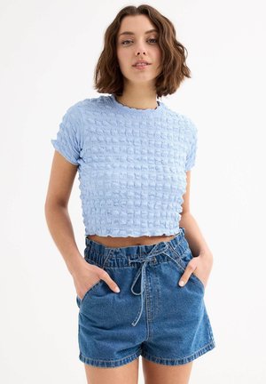Top corto de color azul claro con una superficie texturada y ondulada, mangas cortas y un escote con bordes en forma de ola, combinado con pantalones cortos de mezclilla con cintura elástica.