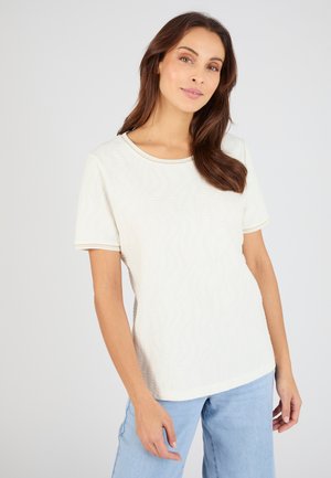 Femme aux longs cheveux bruns portant un haut blanc texturé à manches courtes et un jean bleu clair, debout devant un fond clair uni.