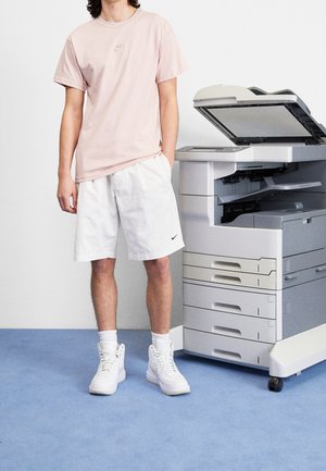 Lyserød bomulds-T-shirt med lille logo, hvide shorts og hvide høje sneakers; grå kontorkopimaskine i baggrunden.