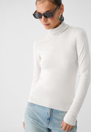 Maglione bianco a collo alto con un design aderente e orlo a coste. Abbinato a jeans azzurri. Nessun motivo o logo visibile. Texture liscia.