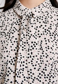 Blouse blanche à carreaux noirs irréguliers et petits points roses, avec un col à nœud papillon noué, portée par une personne.