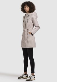 Parka beige clair avec капuche à cordon, taille ceinturée et deux poches avant ; associée à des leggings noirs et des baskets.