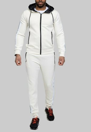Survêtement blanc avec une capuche noire, un devant zippé et des rayures latérales. Associé à des baskets noires avec des lacets orange.