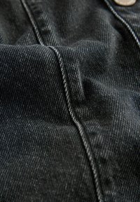Mörk denimtyg med en slät textur, med kontrasterande ljusa sömmar och en synlig knapppunktion. Materialet är något skrynkligt.