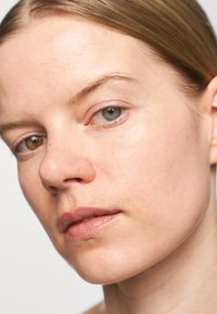 Close-up of a face with smooth skin and minimal makeup. Features include one blue and one brown eye, light brown hair, and a neutral background.
