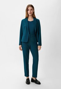 Turkusowa marynarka i dopasowane slim-fit spodnie, wykonane z gładkiego materiału. W zestawieniu z czarnymi loafersami, na neutralnym szarym tle.