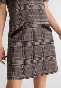 Robe à carreaux en tissu tissé avec manches courtes, dotée de poches noires et d'un ourlet au-dessus du genou. Les couleurs incluent des tons beige, rouge et bleu.