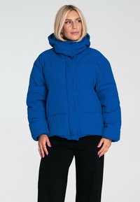 Niebieska kurtka puchowa z kapturem, posiadająca pikowany wzór, zatrzaski oraz elastyczne mankiety, zestawiona z czarnymi spodniami.