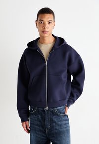 Sweat à fermeture éclair bleu marine en mélange de coton avec une poche avant, capuche et poignets côtelés. Porté par-dessus une chemise beige et associé à un jean bleu.