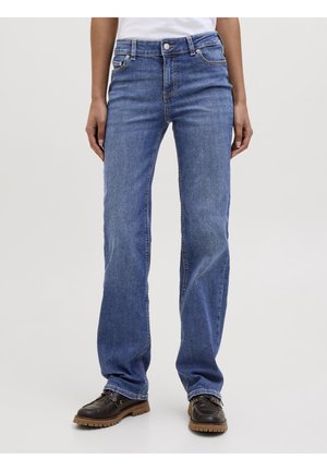 Personne portant un jean droit bleu moyen et des chaussures à lacets marron foncé, debout devant un fond gris clair.