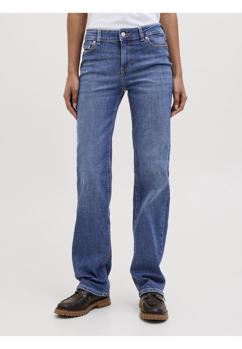 Personne portant un jean droit bleu moyen et des chaussures à lacets marron foncé, debout devant un fond gris clair.