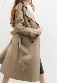 Trench-coat beige avec des boutons croisés, une taille ceinturée et des poches latérales. Texture lisse, longueur jusqu'aux genoux, avec un col classique.