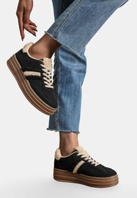 Baskets noires avec un col en fourrure beige texturé, lacets crème et rayures noires et beiges duales. Elles présentent une semelle épaisse en gomme et un jean bleu effiloché.