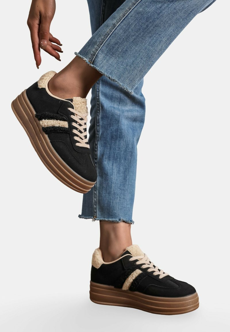 Baskets noires avec un col en fourrure beige texturé, lacets crème et rayures noires et beiges duales. Elles présentent une semelle épaisse en gomme et un jean bleu effiloché.