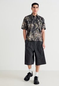 Chemise à manches courtes à imprimé camouflage avec des nuances de vert, marron et noir. Un short à carreaux noir et des baskets noires avec des chaussettes blanches complètent la tenue.