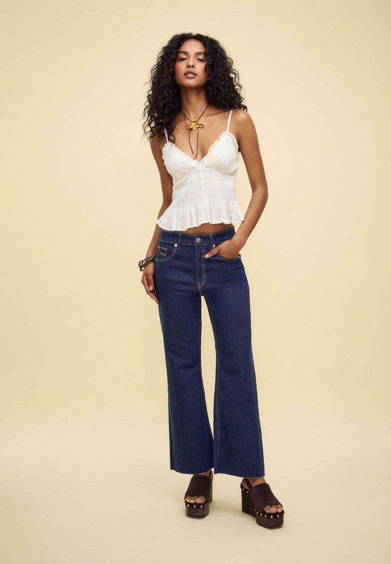 Leichtes weißes Peplum-Top mit Spaghettiträgern und Rüschensaum, kombiniert mit dunkelblauen Schlagjeans und braunen Plattformclogs.
