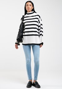 Suéter de punto a rayas en blanco y negro con cuello alto, combinado con jeans de color azul claro y mocasines negros. La modelo sostiene un bolso negro.