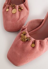Ballerines en daim rose avec bords froncés et breloques dorées décoratives comprenant une étoile, un disque, un nœud et une petite sphère.