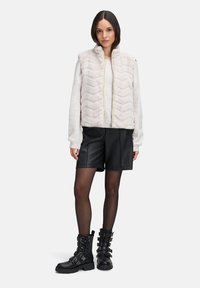 Hellrosa Weste mit Zickzack-Muster, Reißverschluss und gelben Akzenten, kombiniert mit schwarzen Leder-Shorts und schwarzen Stiefeletten mit Schnallen.