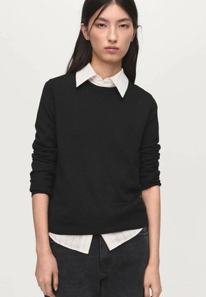 Pull en maille noir avec un col rond, porté sur une chemise blanche à col avec des rayures. Il dispose de manches longues et d'une coupe décontractée.